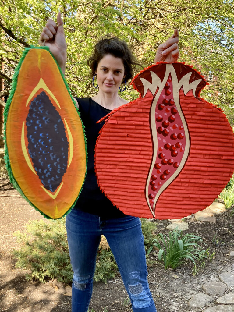 Artist Ashley Cecil holds two large piñatas shaped like fruit