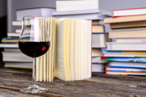 book and wine glass in front of piles of different books on wooden table