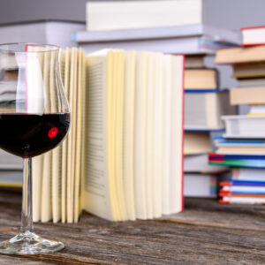book and wine glass in front of piles of different books on wooden table