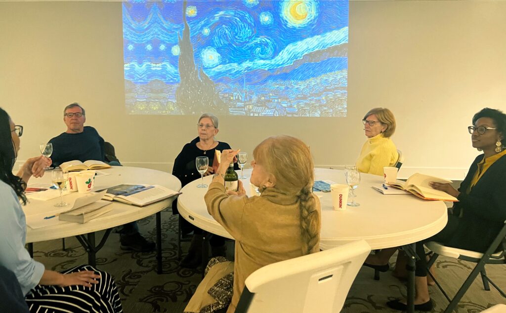 Six members of a book club sit at two round tables with an image of a Vincent van Gogh painting projected in the background.