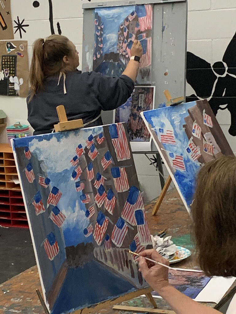 A woman paints a United States flag on a canvas on an easel and two other U.S. flags are being painting on canvases in the foreground.