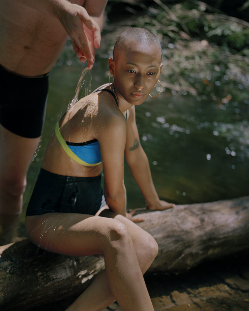 Woman sits on tree branch by creek as someone pours water on her back