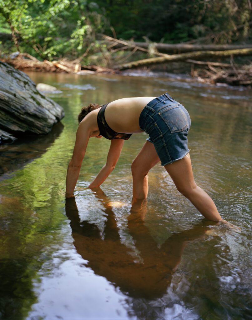 A woman in top and shorts bends over to pick something up in a creek or stream