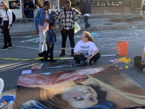 Artist talks with festival goers as she creates chalk image of woman on pavement