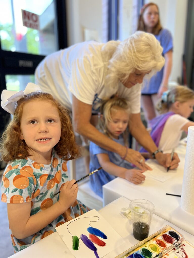 A toddler smiles at the camera during an art project while the teacher helps another toddler in the background