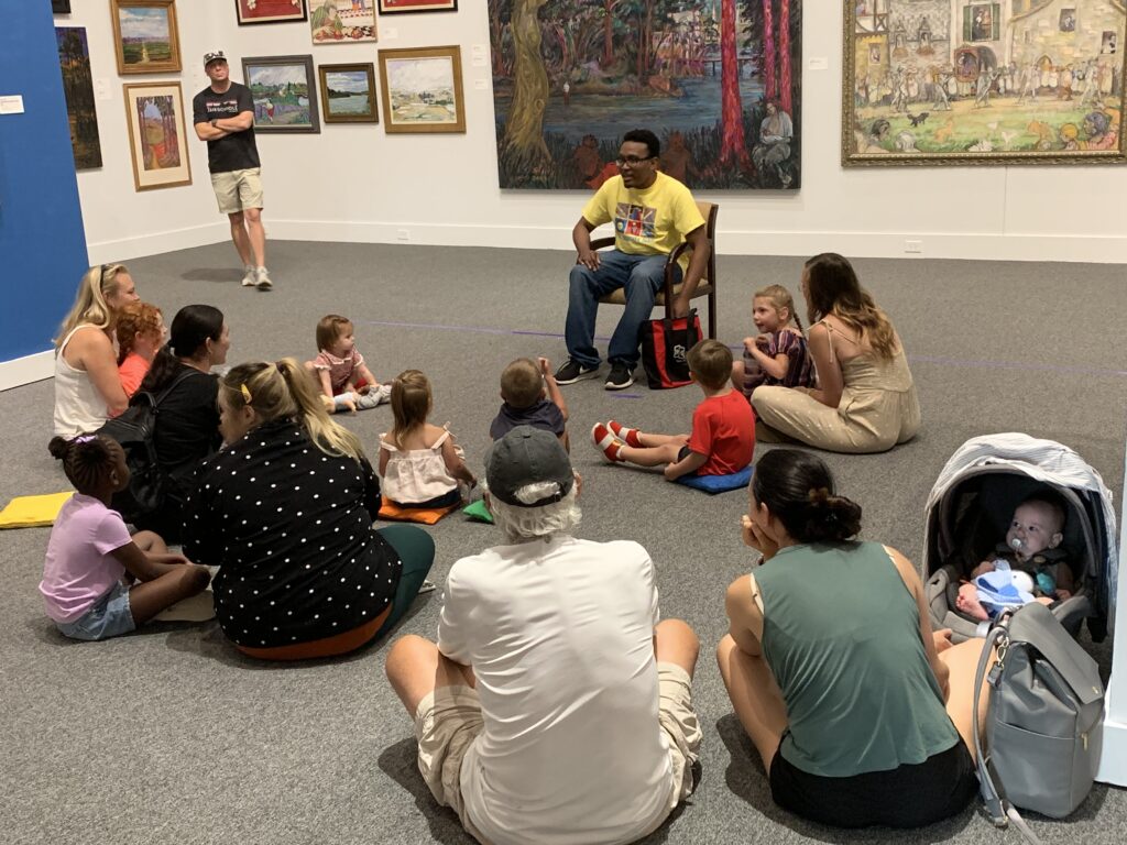 Parents and children are gathered in front of a storyteller in an art gallery