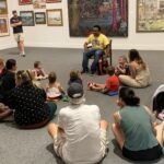 Parents and children are gathered in front of a storyteller in an art gallery