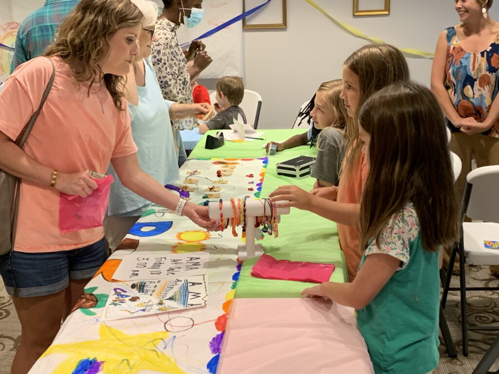 A woman looks at an item that girls have for sale at a pop-up market at art camp
