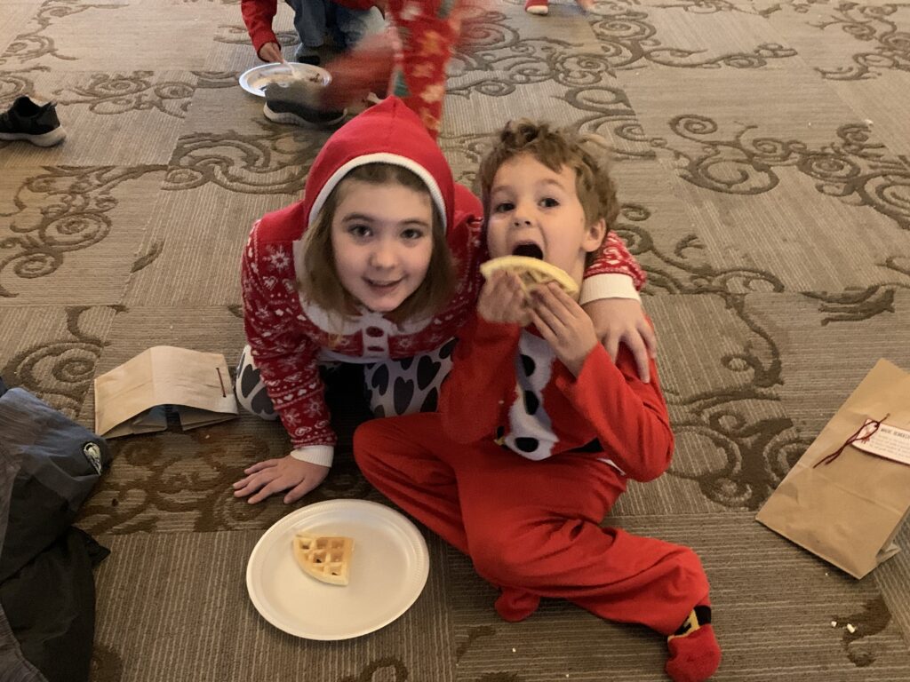 Two kids dressed in holiday pajamas pose as one takes a bite from a waffle