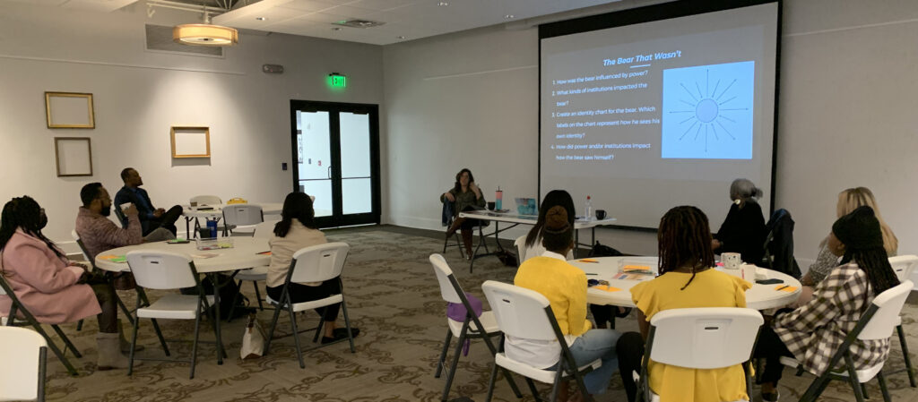 Students and educators sit at round tables with two facilitators at the front. Also at front is a large projection screen used in the Courageous Conversations About Race session.