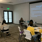 Students and educators sit at round tables with two facilitators at the front. Also at front is a large projection screen used in the Courageous Conversations About Race session.