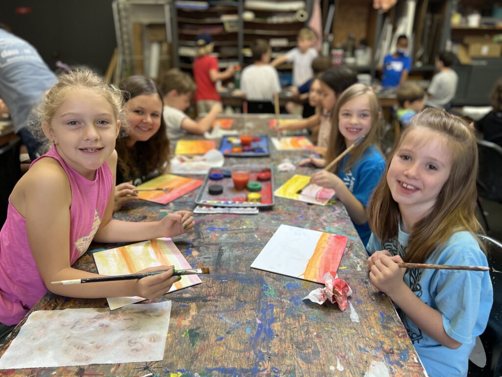 Four campers at a table smile while working on an art project; other campers in grades K-5th grade can be seen in the background.