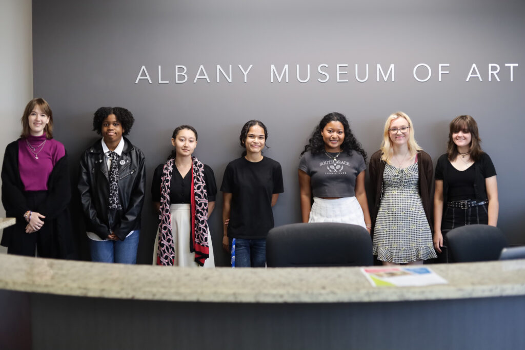 Seven teenagers stand in front of the Albany Museum of Art sign in the museum lobby.