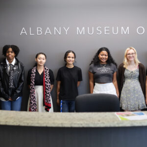 Seven teenagers stand in front of the Albany Museum of Art sign in the museum lobby.