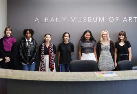 Seven teenagers stand in front of the Albany Museum of Art sign in the museum lobby.