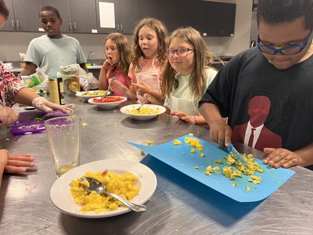 A camper prepares food on a large stainless steel prep table with other campers looking on.