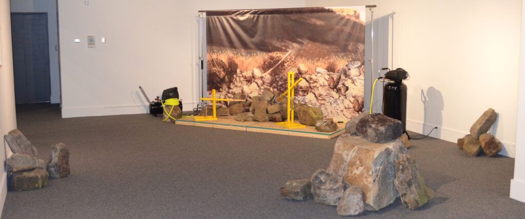 Mechanical sledge hammers break rocks in front of a background of a photograph Gustauve Courbet’s 1849 work, “The Stonebreakers.”