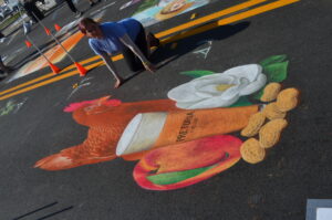 Artist poses with 3D chalk image of chicken, beer, peanuts, peach and magnolia flower