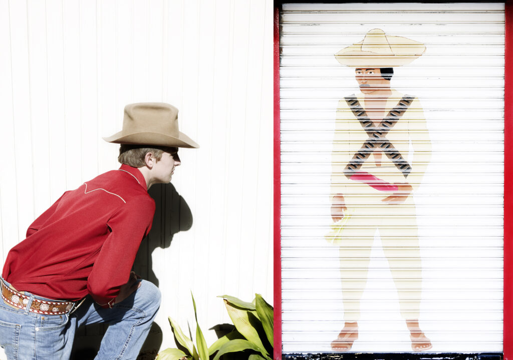 Boy in red shirt and cowboy hat poses looking at image of Mexican character painting onto slats
