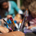 Kid is holding a metal container full of paintbrushes at outdoor arts event