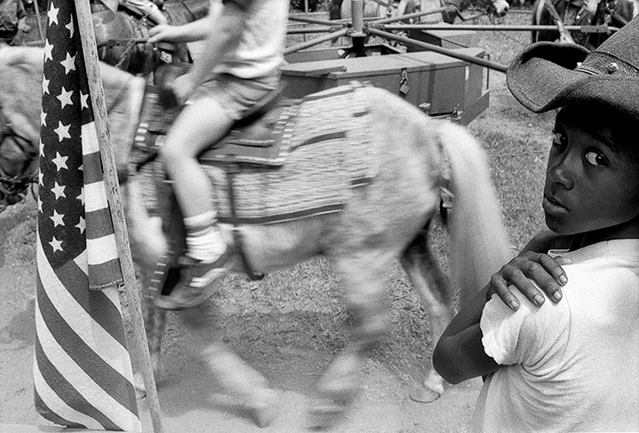 A child in a cowboy hat looks at the camera as another child rides by on a horse at a fair in Gay, Georgia
